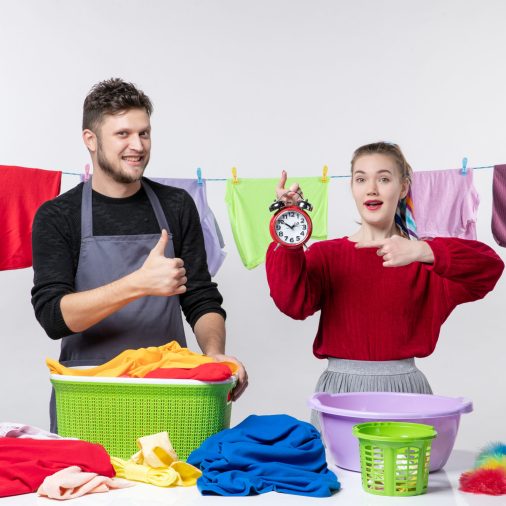 front-view-man-making-thumb-up-sign-his-wife-pointing-alarm-clock-standing-table-laundry-baskets-washing-stuffs-table